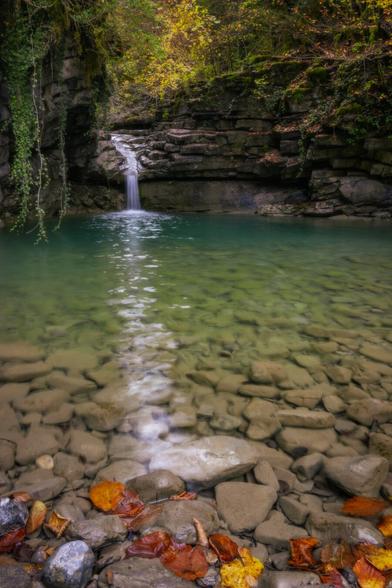 You can see a small natural pond of water. In the foreground, stones and leaves and in the background some autumn vegetation and a small waterfall.