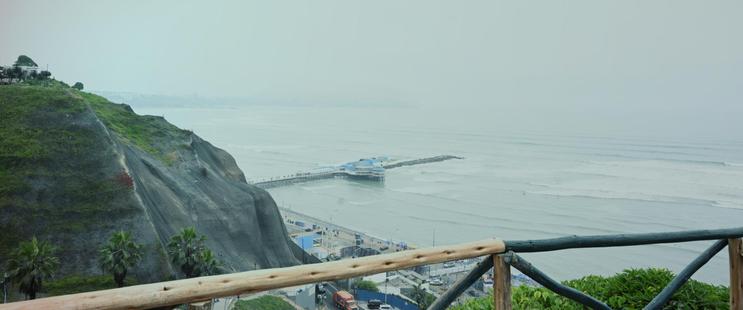 A view of a gray, misty ocean from a high place. A rail of manzanita (?) wood is in the foreground to suggest where the photographer stands. In the distance is a long pier into the water with a blue party boat parked in front. Photo by Peachfront. Lima, Peru.