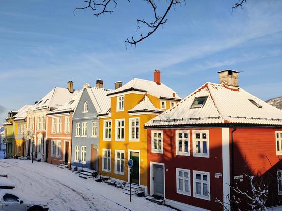 A photograph of a row of small colourful residential houses with a light snow cover. Blue sky behind and some snowed in cars in front.