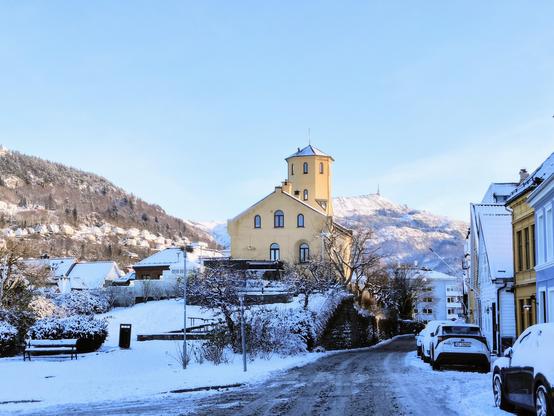 A photograph of an open plaza in a snow covered city. The 17th century city guard building Corps de Garde is shown in center. The snow-covered mountains Fløyen (left) and Ulriken (right) are shown in the background against a light blue sky.