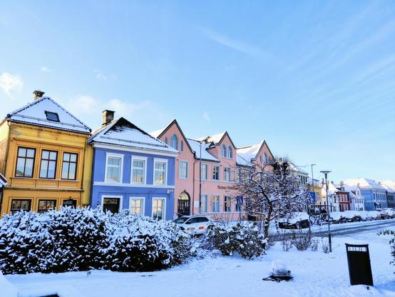 A photograph of a row of small colourful residential houses with a light snow cover. Blue sky with light clouds behind and snow-covered bushes and a small park in front.