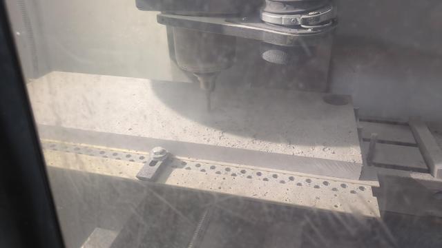 A thick grey rectangle clamped inside a vertical mill, photographed from behind scuffed safety glass. Half inch endmill is cutting into it.