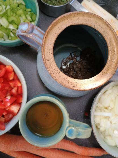 A cup of tea. A teapot with just the tea leaves. Surrounded by bowls of diced red pepper, diced onion, diced celery, and a handful of carrots