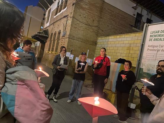 Un grupo de personas jóvenes y de distintos géneros, algunos con banderas trans, de pie en un círculo al lado de un edificio de ladrillo. Llevan velitas en las manos, con un cartón circular para recoger la cera en los colores de la bandera trans. Una sola persona tiene un papel en vez de una velita, como si fuera a leer un manifiesto. Se ve parte del cartel informativo de la antigua cárcel.