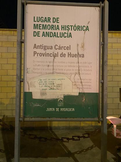 Cartel informativo de la antigua cárcel de Huelva. Señala: lugar de memoria histórica de Andalucía. Antigua cárcel provincial de Huelva. Debajo hay texto ilegible por la falta de luz, está roto, y el espacio en blanco de ese roto está ocupado por un grafiti de rotulador.