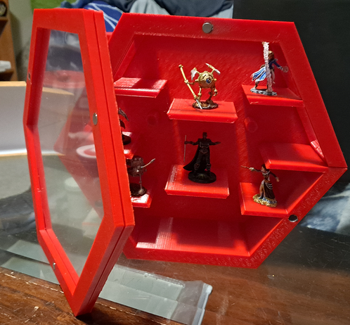 A hexagonal display case in red plastic with clear plastic sheet inserts, with the front panel taken off and positioned beside it to show the magnetic closures, holds a selection of tabletop RPG miniatures.