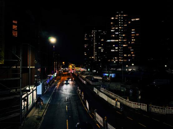 Color photo of a nighttime urban scene. A partially lit street lined with construction barriers and illuminated shop windows on the left. Two persons carrying bags walks along the sidewalk under scaffolding, while two cars drives down the wet road reflecting light. In the background, high-rise apartment buildings with scattered lit windows rise against the dark sky. (Photo by me, CC-BY-4.0)