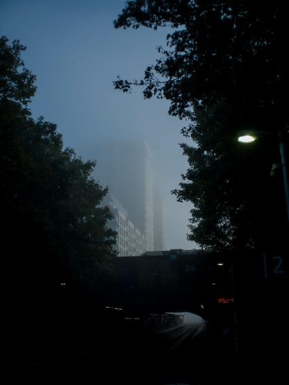 Photo in which a tall white apartment building is partially visible through mist, fading away towards the top. Framing it are silhouettes of nearby trees on either side and a built-upon bridge below it. Beneath the bridge a pair of rail tracks run past platforms into nothingness.