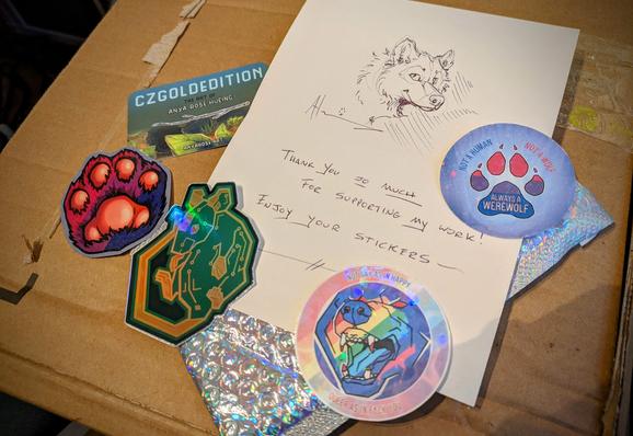 Photo of an open card laid out with stickers, a business card, and a sparkly bubble mailer. The card has a doodle of a wolf in it and says: "Thank you so much for supporting my work! Enjoy your stickers~"