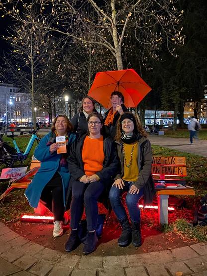 Bürgermeisterin Martina Wild, Stadträtinnen Pia, Haertinger, Christl Kamm, Landtagsabgeordnete Stephie Schuhknecht und ich auf einer orangen Bank am Königsplatz im Auto. Auf der Bank steht rechts: "kein Platz für Gewalt an Frauen". Viele tragen orange Kleidung, ein oranger Regenschirm ist auch da - Zeichen für den "Orange Day" als Wiedererkennungsmerkmal für den 25. November, Tag gegen Gewalt an Frauen. Es hat eine dämmerige Stimmung.