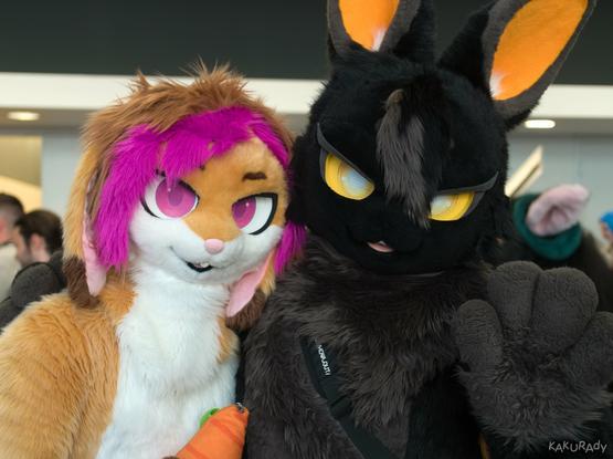 A brown rabbit fursuiter with white belly and magenta highlights, holding the shoulder of a black rabbit fursuiter with dark grey belly and yellow and orange highlights, who waves to the viewer with their left hand.