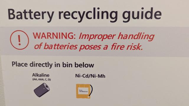 Battery recycling guide poster on wall, with a warning message set in a mechanically skewed oblique text with improper kerning.