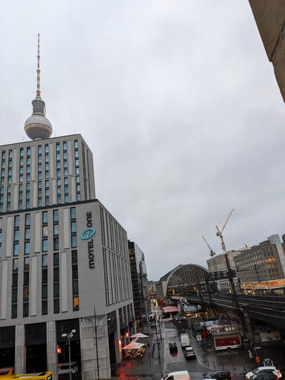 Das Bild zeigt eine Stadtlandschaft an einem bewölkten Tag. Im Vordergrund steht ein modernes, hellgraues Hotelgebäude mit dem Schriftzug "Motel One" an der Seite.  Im Hintergrund ist die Berliner Fernsehturm sichtbar, sowie ein Bahnhof mit Gleisen und ein Baukran. Die Straße vor dem Hotel ist nass und es sind einige Autos zu sehen.  Der Gesamteindruck ist der einer modernen Stadt mit einem Mix aus alten und neuen Gebäuden an einem eher trüben Tag.

Wie im Text geschrieben handelt es sich beim Bahnhof im Hintergrund um den Bhf Berlin Alexanderplatz.