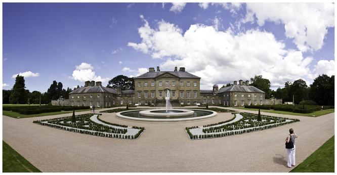 A grand, symmetrical stone mansion surrounded by landscaped gardens and a central fountain. The sky is partly cloudy, and a person is seen walking in the garden.