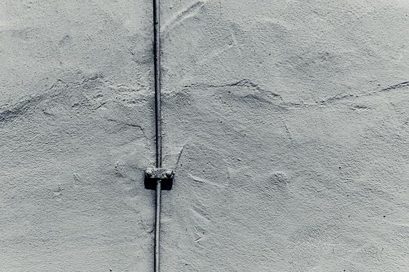 Das Foto zeigt eine minimalistische Komposition einer verputzten Wand in einem matten, monochromen Farbton, vermutlich Grau oder Blau-Grau. Die Oberfläche der Wand weist eine raue Struktur mit feinen Rissen und Unebenheiten auf. In der Bildmitte verläuft vertikal ein dünnes Metallrohr, das mit einer kleinen, rechteckigen Halterung befestigt ist. Das Bild vermittelt eine industrielle und kühle Ästhetik durch die Betonung von Einfachheit, Textur und geometrischer Klarheit.