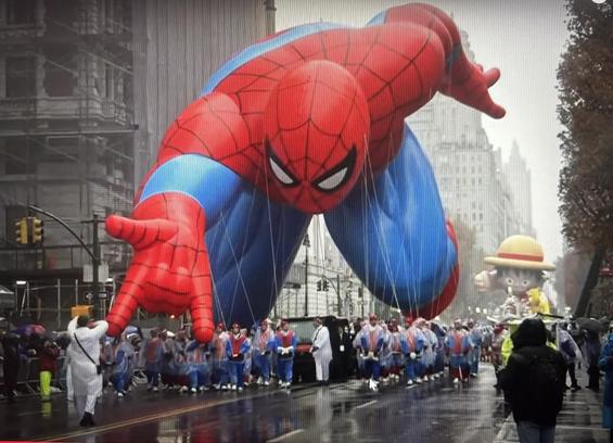The Thanksgiving parade in NYC. Spider-Man float in the foreground, other character float way behind up the street. Both balloons are close to the ground, with Spider-Man’s knee almost touching the pavement. The leaves on the trees are brown and the sky is grey, the crowd has umbrellas.