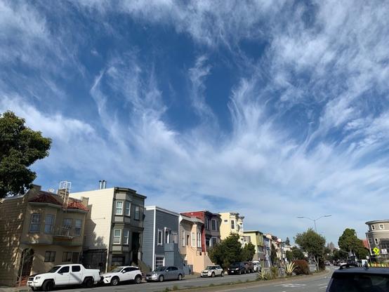 A photo of some clouds over some homes in San Francisco. The sky is a beautiful bright blue filled with large patches of thin, stringy clouds on a sunny day.