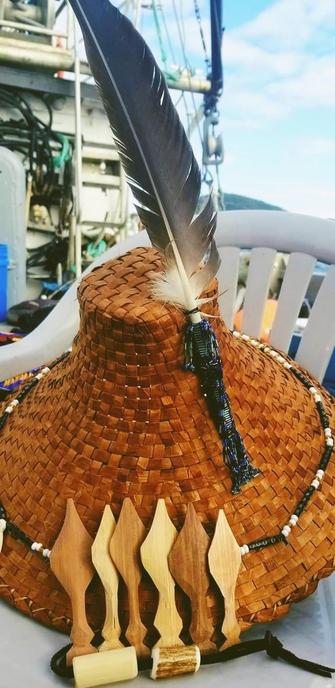 A cedar woven hat sits on a surface, featuring a prominent eagle feather attached at the top, with beading around the brim & wrapped around eagle feather tip.
Several carved cedar paddles of varying shapes and sizes are arranged in front of the hat. The background includes hints of a fishing boat environment with ropes and containers.
