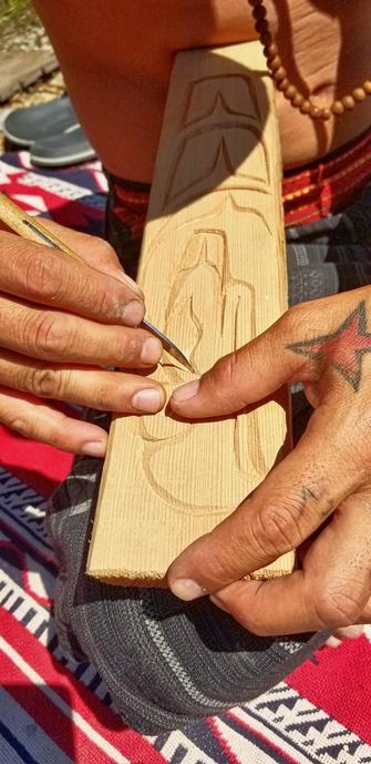 A close-up image of a shirt-less man, wearing a wood beaded necklace and carving a detailed Coast Salish Indigenous design into a small wooden cedar plank. The artist uses a small carving tool while holding the wood steady with their fingers. Their hands show signs of experience, with a tattooed red star visible on one hand. The background is outdoors, in an open natural area.