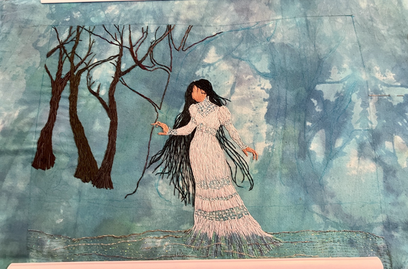 An embroidered panel of La Llorona standing in the edge of the Rio Grande, with twisting mesquite trees being worked behind her.