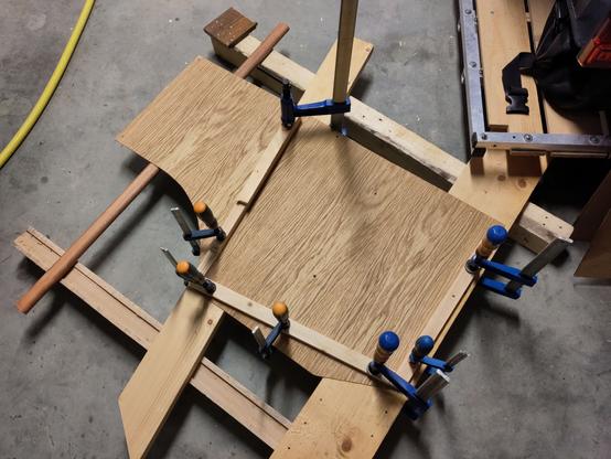 Photo d'une des deux portes en cours de collage. Elle est constituée de deux plaques de placage bois collées l'une sur l'autre (c'était l'étape précédente pour laquelle je n'ai pas de photo), et ici on voit la porte posée sur des planches, elles-mêmes posées sur le sol, avec des tasseaux qui forment un "U" qui servira à rigidifier la porte, et sont maintenus par des serres-joint le temps que la colle sèche.