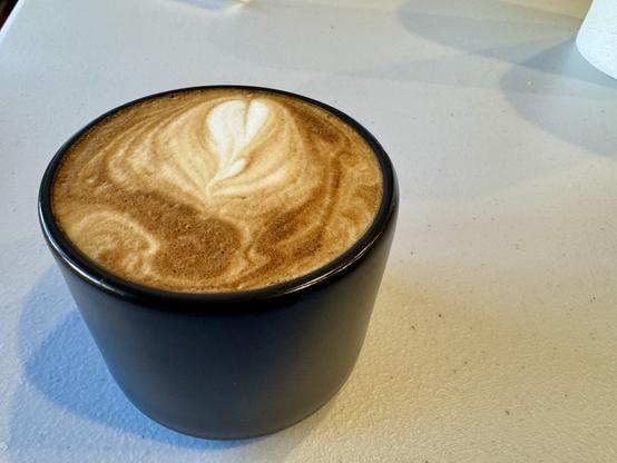 Small black coffee mug filled with a cappuccino siting on a grey white textured table