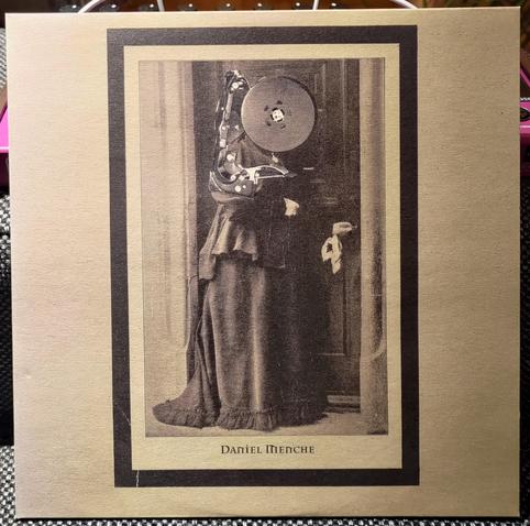 Light brown 10" cover with a vertical framed image. Old black and white photo of lady, but with some sort of device over her face and torso. Looks like a tape reel or something.
"Daniel Menche" under the image