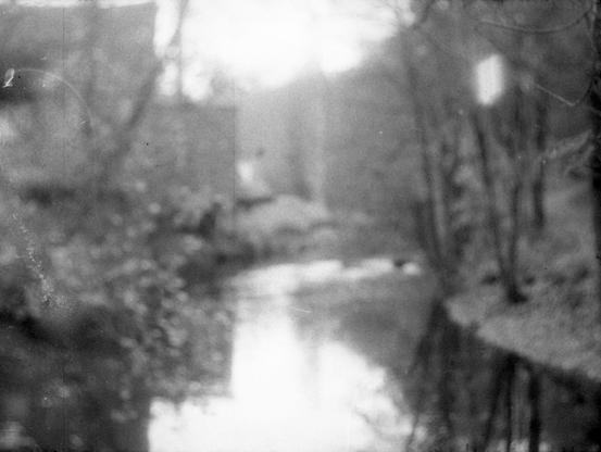 blurry black and white image with river in the middle, trees on the benches and a factory in the background