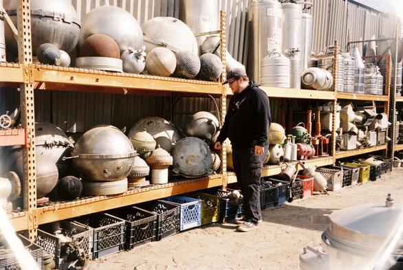 A guy with beard, glasses and cap infront of a rack of round metall balls, that look like watermines.