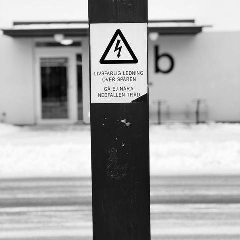 The image shows a black metal pole with a warning sign attached to it. The sign has a triangular warning symbol with a lightning bolt inside, indicating danger. The text on the sign is in Swedish and reads: 

**"LIVSFARLIG LEDNING ÖVER SPÅREN"**  
**"GÅ EJ NÄRA NEDFALLEN TRÅD"**

This translates to: 

**"Lethal power lines over the tracks"**  
**"Do not go near fallen wires"**

The background features a snowy ground and a blurred building, possibly a public facility or station. The image is in black and white.