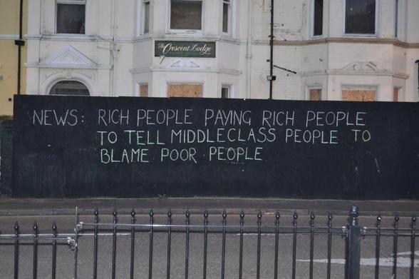 Street art on a black wall outside a boarded-up building with the text: 'NEWS: RICH PEOPLE PAYING RICH PEOPLE TO TELL MIDDLECLASS PEOPLE TO BLAME POOR PEOPLE.' The graffiti, written in simple white chalk-style lettering, critiques societal and economic dynamics. The location appears to be in an urban area, with a fence in the foreground and a sign reading 'The Crescent Lodge' visible on the building behind the wall.