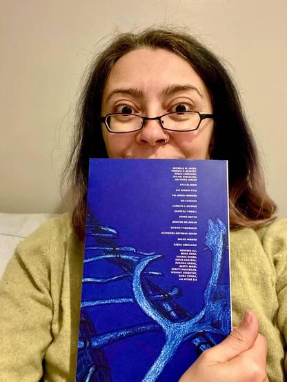 A person is holding a blue book cover featuring an abstract design. They have long hair and are wearing glasses. The person appears to have a surprised or joyful expression.