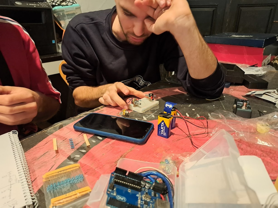 Una mesa roja está llena de componentes electrónicos, alambres y herramientas. Dos personas trabajan juntas en un proyecto de electrónica. Una persona se inclina sobre un prototipo de circuito pequeño conectado a una batería. Se ve frustrado y se frota la frente con la mano. Alrededor del prototipo hay resistencias, un microcontrolador y otros componentes electrónicos. La otra persona está sentada a su lado, observando el trabajo. Sobre la mesa hay un cuaderno y un teléfono móvil.  La escena sugiere un taller o un entorno de trabajo colaborativo relacionado con la electrónica.