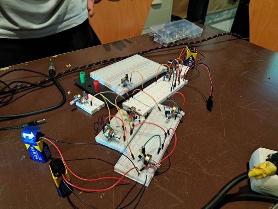 Una mesa de madera oscura muestra varios prototipos de circuitos electrónicos.  Hay varias placas de pruebas con componentes electrónicos como resistencias, potenciómetros y cables de colores conectados. Las placas están interconectadas entre sí.  Dos baterías de 9V son visibles, junto con un cable de audio y una caja de componentes electrónicos.  Se ve la parte inferior de una persona en segundo plano. La escena sugiere un proyecto de electrónica en progreso.