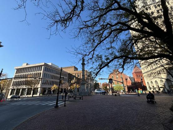 A city square.  Open space and a tree in shadow in the foreground.  Various buildings in sunlight in the background.