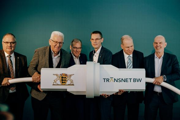 Ministerpräsident Winfried Kretschmann (zweiter von links) hält zusammen mit fünf weiteren Männern zwei überdimensionierte weißen Stromstecker, die zusammengesteckt wurden, um den Konverter symbolisch in Betrieb zu nehmen.  Auf den Steckern ist das Baden-Württemberg-Wappen und das TransnetBW-Logo zu sehen. © Staatsministerium BW | Ilkay Karakurt