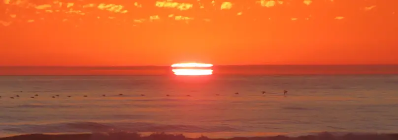 A photograph a sunset over the Pacific Ocean. The sun appears to be cut in half by a cloud. A line of pelicans flies over the water.