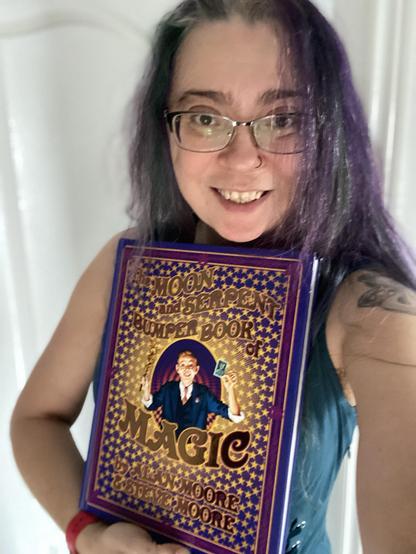 A person is smiling while holding a large hardcover book titled "The Moon and Serpent Bumper Book of Magic" by Alan Moore and Steve Moore. It is purple and metallic gold and heavy enough to use as a murder weapon.