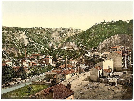 The image depicts a historical photograph showcasing an urban landscape with industrial and residential elements. The scene is set in Fiume, Zwirthal during the Austro-Hungarian Empire era between 1890 to around 1900. In this picturesque view, we observe numerous buildings clustered together along winding streets that meander through a mix of greenery and rocky terrain.
In the foreground, there's an abundance of red-roofed houses tightly packed alongside a river flowing from left to right; its waters are a striking blue-green shade contrasting with the earthy tones surrounding it. To the upper regions stands imposing structures such as chimneys emitting smoke, hinting at industrial activity within this area.
The midground is dominated by more densely populated buildings that blend into one another creating an impression of small town life during late 19th-century Europe's Industrial Revolution. Behind these dwellings looms a hillside with vegetation providing some natural relief to the urban sprawl above, while higher up sits what seems like fortifications or residential structures atop this ridge.
Towards the background, we notice more industrial elements such as larger buildings possibly serving commercial purposes and towering over smaller edifices in their vicinity. The sky is clear with no visible clouds suggesting a calm day free from adverse weather conditions typical of that period's European climate. Overall, it [...]