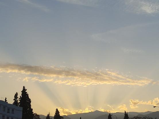 Se ven una casas y al fondo unas montañas y en el cielo la luz es de amanecer y se ven luces crepusculares como haces entre unas cuanta nubes, dando al cielo un toque naranja