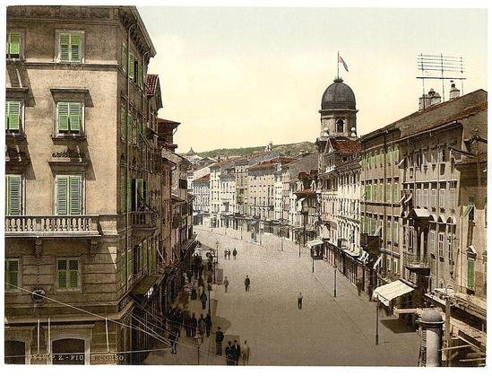 A vintage photograph depicting a bustling street in Fiume, Croatia during the Austro-Hungarian Empire. The scene captures an array of historic buildings with green shutters and decorative facades lining both sides of the wide thoroughfare. People are seen walking along the sidewalks or crossing the street at various points. A prominent dome-topped building rises above others towards a hazy skyline, suggesting it may be a notable landmark in this urban landscape.