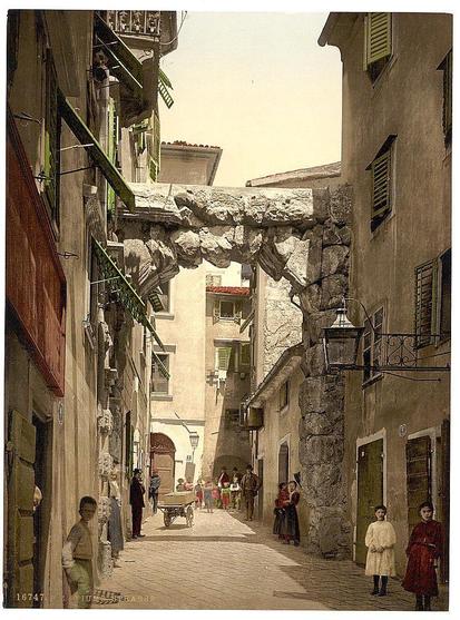 This image depicts a picturesque street in Fiume, Croatia during the Austro-Hungarian Empire era. The architecture features old stone buildings with colorful shutters on their windows and doors. A prominent feature is an ancient archway constructed from weathered stones that spans over the narrow road, connecting two sides of the alleyway.
Several people are seen strolling along this charming street, dressed in period clothing indicative of late 19th or early 20th-century fashion. Men wear suits and hats, while women sport long dresses with various headwear styles. The overall ambiance suggests a bustling yet quaint urban environment from that historical time frame.