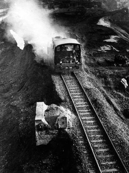 The black and white photograph depicts a steam locomotive operating on railway tracks. The train appears to be shunting, maneuvering goods around or into the vicinity of what seems like an industrial site with visible coal heaps and excavation activity in the background.

In terms of visual elements:

- A central focus is placed on a vintage steam engine emitting smoke from its chimney.
- We see workers wearing hats operating within the train's cab. The number '351' is marked on the side, likely denoting the locomotive's identification or model.
- Tracks are laid out with stones and gravel between them; tracks extend into the distance but don't clearly show where they lead to.
- There appears to be a significant amount of excavation happening in front of the train. The earth has been dug up, exposing layers of rock and soil.

This image captures an industrial scene likely from mid-20th century given its monochromatic nature and style of locomotive design. It conveys an atmosphere indicative of heavy industry during that era, with a focus on labor-intensive processes such as coal mining or transportation around the worksite.