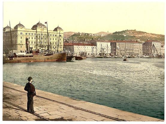 A vintage photo featuring a man standing on a quay, overlooking the harbor of Fiume (now known as Rijeka) in Croatia. He is dressed in period attire with a hat and suit jacket indicative of late 19th or early 20th-century fashion. Behind him lies a large body of water reflecting sunlight with several sailing ships anchored near the dock, some smaller boats scattered around, and various buildings lining the waterfront including what appears to be an imposing port building with classical architectural features such as domes and columns.

In the background on land, other buildings can be seen in shades of cream, red, and white. Further back atop a hill or elevated terrain sits a fort-like structure under clear skies, suggesting Fiume's strategic importance during its time. The image is tinged with an earthy tone typical of early photography techniques such as photochrom printing from the late 19th to mid-20th century.

The bottom right corner bears the inscription "8416-FZ" and a date or initials "F. E." which could signify its cataloging in archives, likely pertaining to Austrian Empire's cultural records during that era.