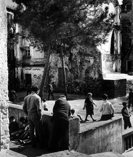 The image is a black and white photograph depicting an urban scene. In the foreground, several adults are gathered around children who appear to be sitting on steps or ledges within what appears to be an old courtyard area of a dilapidated building with exposed brickwork.

There's graffiti visible in various places including "CREA GIOCONDO" which is partially obscured and another message that seems like it could read as "MOLTO," suggesting the presence of people who have defaced or marked their surroundings. Some individuals are standing while others sit, creating a relaxed but casual atmosphere with some engaged in conversation.

The background features tall buildings made from stone and brick, indicating an older part of town possibly undergoing decay or disrepair. The foliage is sparse at best; there's one large tree that provides little to no shade for the area where people are congregating.

Overall, this photo captures a slice-of-life moment within what appears to be a rundown neighborhood in Europe during perhaps late 20th-century times given its style and urban decay characteristics.