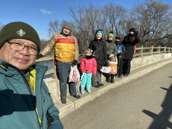 A group of adults and kids bundled up for cold weather standing along the edge of a bridge while holding the railing. They are squinting and smiling into the camera. Leafless trees are in the background.