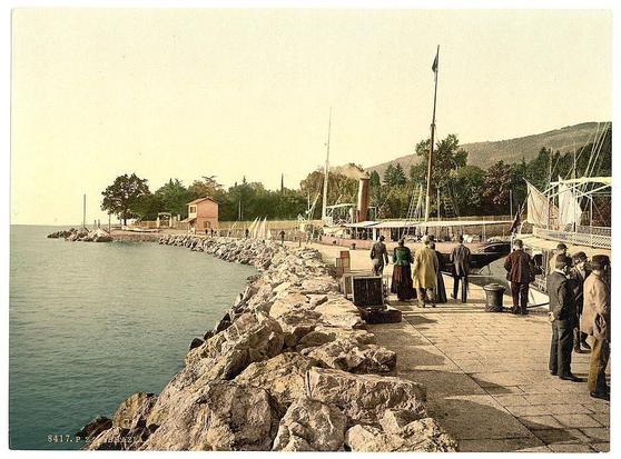 The image depicts a vintage scene at a coastal harbor, likely in the late 19th or early 20th century. A group of individuals is gathered on a stone-paved walkway beside calm waters with boats moored nearby. Some people appear to be conversing while others are walking along the path. In the background stands an old-fashioned steamboat docked at the pier, surrounded by lush greenery and trees. The shoreline features large rocks leading up to what seems like residential or commercial buildings in close proximity to the water's edge. A flag is hoisted atop a pole near the center of the image, suggesting some form of official presence or establishment nearby. Overall, it captures an era where maritime activity was common along coastlines and harbors were bustling centers for trade, transportation, and social gatherings.