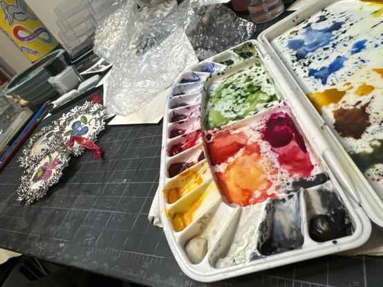 The absolute chaos of my studio desk. A watercolor palette in the foreground, some finished watercolor ornaments in the background, some bubble wrap, paint brushes, and god knows what else.