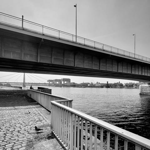 das quadratische schwarzweißfoto zeigt den rhein in köln, unterhalb der deutzer brücke in richtung rheinauhafen.
in der oberen bildhälfte ist klarer himmel zu sehen, ebenso wie ein teil der deutzer brücke von schräg unten.
in der unteren bildhälfte ist das geländer am kennedy-ufer zu sehen. das geländer verläuft in zickzack-form in die rechte untere bildecke. der Boden ist mit steinen gepflastert.
in dem verbleibenden schmalen streifen zwischen brücke oben und rhein unten verläuft der horizont.  zu sehen sind gebäude auf der linken rheinseite wie die kranhäuser. auch die severinsbrücke ist zu sehen.
die komposition von brücke, horizont und handlauf hat ebenfalls eine zickzack-struktur.
