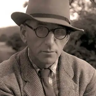 Patrick Kavanagh wearing a trilby style hat, glasses, checked shirt, tie and tweed jacket.